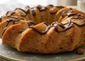 Ciambella alle castagne soffice e dorata servita su un piatto, decorata con una colata di cioccolato fondente e zucchero a velo. Dolce autunnale fatto in casa, ideale per la colazione o la merenda, con castagne e nocciole in evidenza.