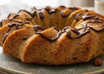 Ciambella alle castagne soffice e dorata servita su un piatto, decorata con una colata di cioccolato fondente e zucchero a velo. Dolce autunnale fatto in casa, ideale per la colazione o la merenda, con castagne e nocciole in evidenza.