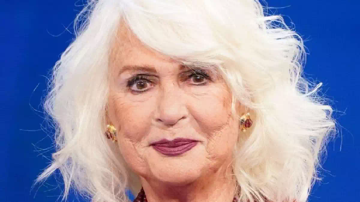 Loretta Goggi in primo piano durante un'apparizione pubblica, con capelli bianchi e sfondo blu. La celebre artista italiana ha annunciato la sua scelta di restare lontana dalla televisione