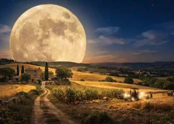 Gigantesca Luna piena del Raccolto che domina il cielo notturno sopra una campagna collinare, con casale rurale, campi dorati appena mietuti, trattore al lavoro e sentiero di campagna illuminato dalla luce lunare. Suggestiva scena autunnale che richiama le antiche tradizioni agricole legate al plenilunio di settembre.