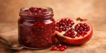 Vasetto di marmellata di melograno aperta senza etichette, accanto a un melograno tagliato a metà su sfondo neutro dai toni caldi, ideale per articoli di cucina naturale e ricette senza zucchero