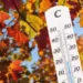 Foglie autunnali dai colori arancioni, gialli e rossi illuminate dal sole con termometro in primo piano che indica temperature miti, simbolo dell'Ottobrata e dell'autunno caldo in Italia.