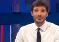 Stefano De Martino in primo piano durante una trasmissione televisiva negli studi Rai, con camicia bianca e cravatta scura. Il conduttore è seduto davanti alle telecamere con alle spalle uno sfondo illuminato da luci blu