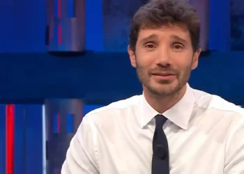 Stefano De Martino in primo piano durante una trasmissione televisiva negli studi Rai, con camicia bianca e cravatta scura. Il conduttore è seduto davanti alle telecamere con alle spalle uno sfondo illuminato da luci blu