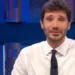 Stefano De Martino in primo piano durante una trasmissione televisiva negli studi Rai, con camicia bianca e cravatta scura. Il conduttore è seduto davanti alle telecamere con alle spalle uno sfondo illuminato da luci blu