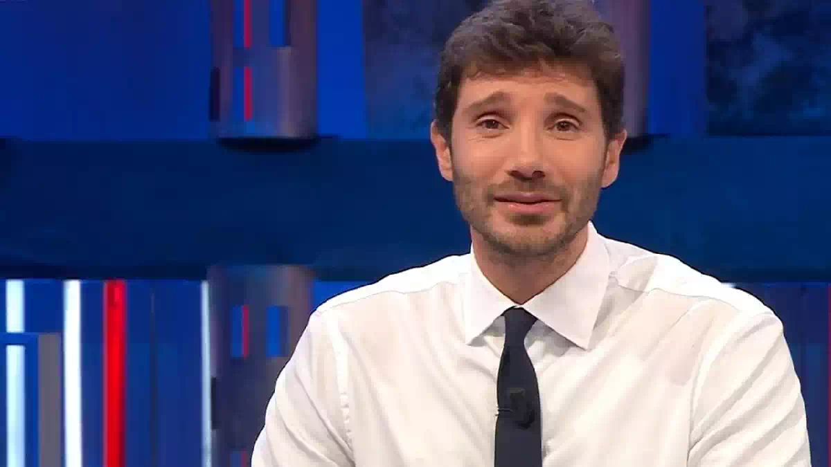 Stefano De Martino in primo piano durante una trasmissione televisiva negli studi Rai, con camicia bianca e cravatta scura. Il conduttore è seduto davanti alle telecamere con alle spalle uno sfondo illuminato da luci blu