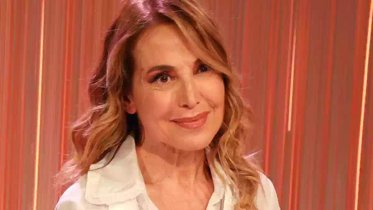 Barbara D’Urso in primo piano durante un evento televisivo, con sfondo illuminato da luci arancioni e rosse