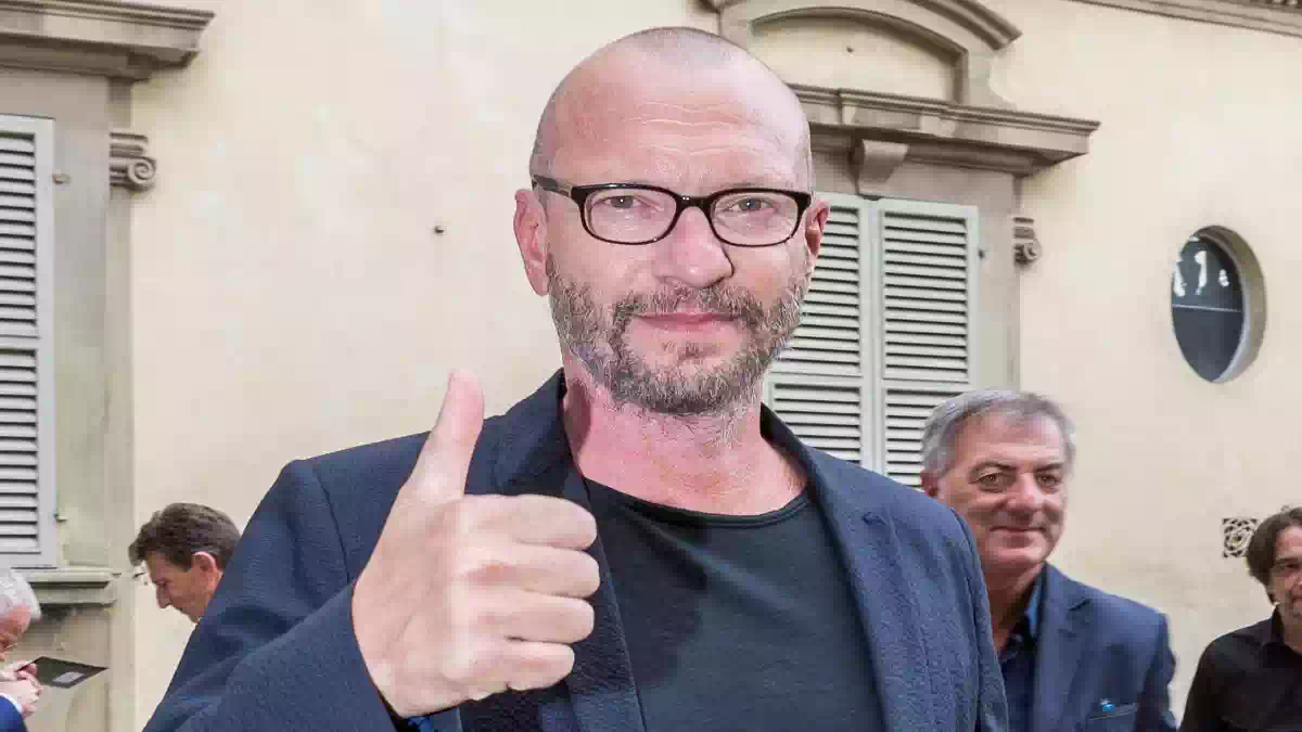 Biagio Antonacci durante una cerimonia pubblica a Rozzano, ritratto in primo piano con il pollice alzato davanti a un edificio della città