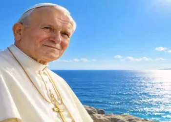 San Giovanni Paolo II in primo piano davanti al mare azzurro e scintillante, con cielo limpido e luce solare intensa all’orizzonte. Ritratto luminoso e realistico del Pontefice in abito bianco