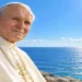 San Giovanni Paolo II in primo piano davanti al mare azzurro e scintillante, con cielo limpido e luce solare intensa all’orizzonte. Ritratto luminoso e realistico del Pontefice in abito bianco