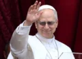 Papa Leone XVI saluta i fedeli con la mano alzata durante un'apparizione pubblica, indossando l'abito bianco e gli occhiali scuri davanti a un sipario rosso