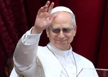 Papa Leone XVI saluta i fedeli con la mano alzata durante un'apparizione pubblica, indossando l'abito bianco e gli occhiali scuri davanti a un sipario rosso