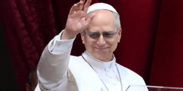 Papa Leone XVI saluta i fedeli con la mano alzata durante un'apparizione pubblica, indossando l'abito bianco e gli occhiali scuri davanti a un sipario rosso