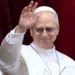 Papa Leone XVI saluta i fedeli con la mano alzata durante un'apparizione pubblica, indossando l'abito bianco e gli occhiali scuri davanti a un sipario rosso