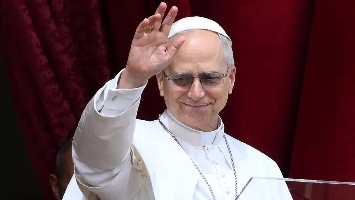 Papa Leone XVI saluta i fedeli con la mano alzata durante un'apparizione pubblica, indossando l'abito bianco e gli occhiali scuri davanti a un sipario rosso