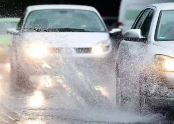 Maltempo con forte pioggia su una strada urbana: automobili in marcia tra pozzanghere e spruzzi d’acqua sull’asfalto bagnato durante un intenso temporale
