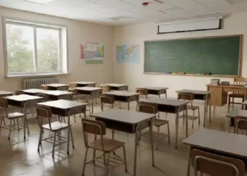 un'aula scolastica vuota, con diversi banchi e sedie in legno disposti in file ordinate. I banchi sono rivolti verso una grande lavagna verde appesa a una parete color crema.