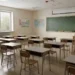 un'aula scolastica vuota, con diversi banchi e sedie in legno disposti in file ordinate. I banchi sono rivolti verso una grande lavagna verde appesa a una parete color crema.